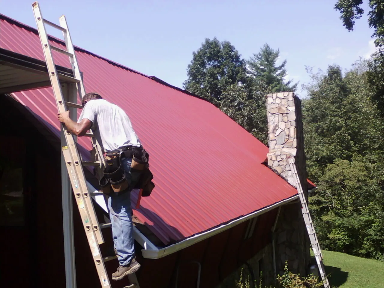 Roofer on a ladder beside a red metal roof and stone chimney — Soffit & Fascia Repair by Roofing Brothers in Peachtree Corners